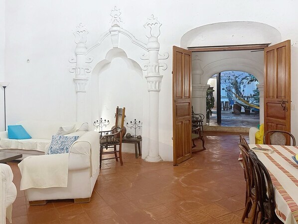 Um quarto com um sofá branco, uma cadeira de madeira, uma mesa de jantar com cadeiras e vista para um jardim através de uma porta aberta.