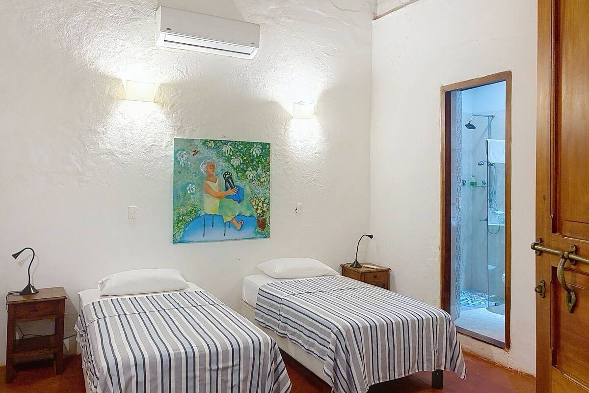 A room with two beds, a painting on the wall, an air conditioner, a wooden door, and a shower area.