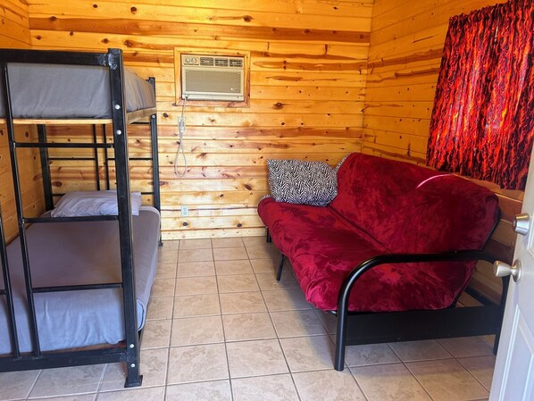 A bunk bed with a red sofa and a patterned pillow, wooden walls, and a window with a curtain. - Sturgis Camping Cabin with Futon and Bunkbed (Sturgis)