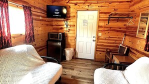 Interior de uma cabana com televisão na parede, uma porta e uma pequena mesa com cadeiras.