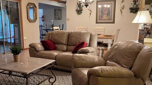 Ein Wohnzimmer mit einem beigefarbenen Ledersofa, einem Couchtisch und einem gemusterten Teppich. Im Hintergrund befindet sich ein Essbereich mit Tisch und Stühlen.