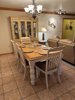 Een eetruimte met een houten tafel gedekt voor vier personen, omringd door witte stoelen. Daarboven hangt een kroonluchter en in de achtergrond is een kast met glazen deuren te zien.
