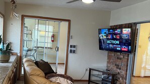 Ein Wohnzimmer mit einem beigefarbenen Sofa, einem Glastisch, einem Deckenventilator und einem Flachbildfernseher, der ein Videospiel zeigt.
