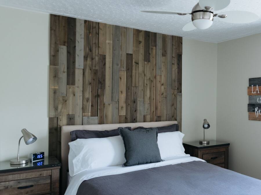 Una camera da letto con testiera in legno, un letto con biancheria da letto bianco e grigio, due comodini con lampade e una ventola a soffitto.
