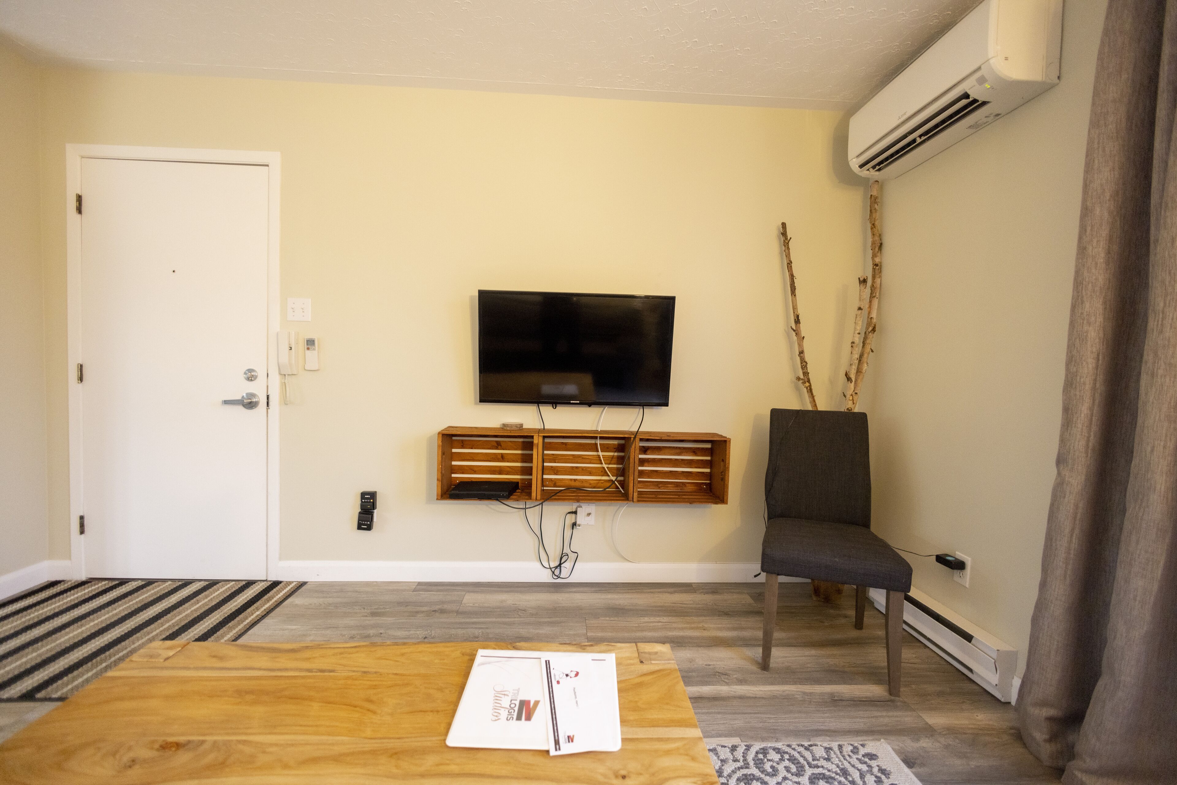 A living room with a flat-screen TV mounted on the wall, a wooden entertainment unit below it, a grey armchair, and a coffee table with a magazine.