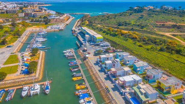 An aerial view of a marina with boats docked, a large industrial building, and residential structures with colorful roofs.