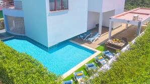 A modern villa with a swimming pool, sun loungers, and a view of a hilly landscape.