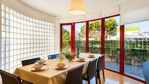 Un coin repas avec une table dressée pour quatre personnes, une grande fenêtre aux montants rouges et une vue sur la verdure à l’extérieur.