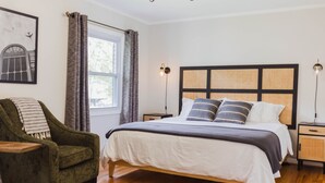 Un dormitorio con cama, una silla, mesita de noche, lámpara y ventana con cortinas.