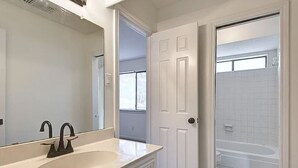 Un baño con un lavamanos blanco, dos llaves, un espejo grande y una puerta que da a la zona de ducha.
