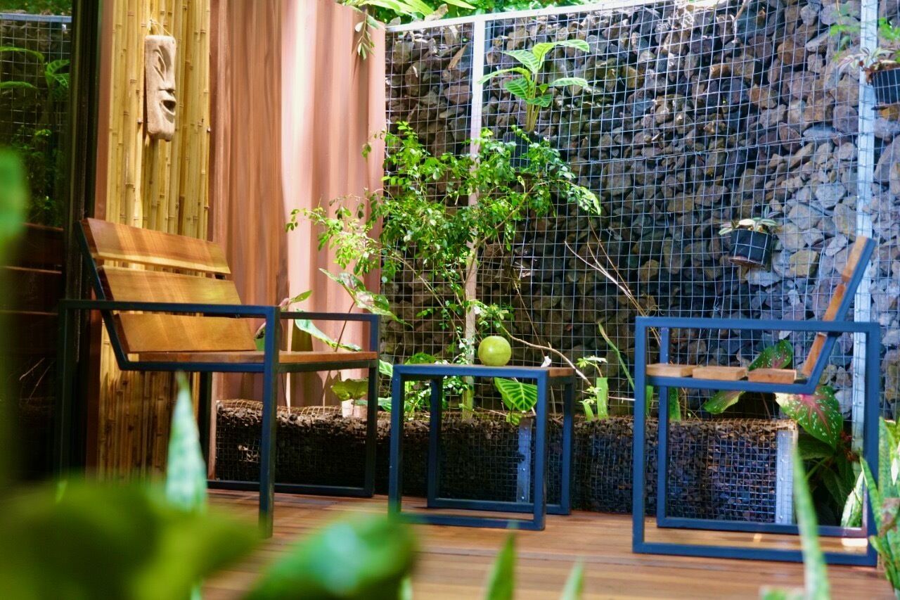 Una terraza de madera con sillas y una mesita, rodeada de plantas exuberantes y un muro de piedra.