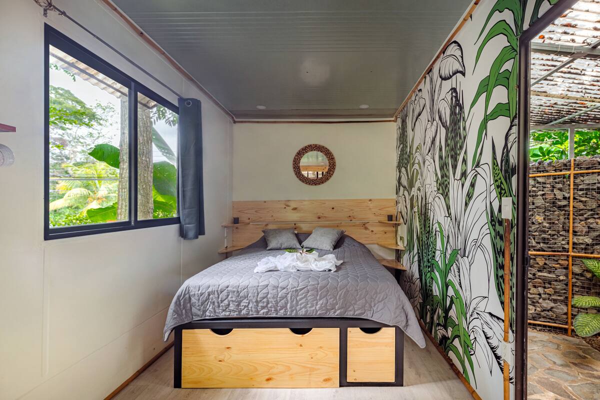 Quarto com cabeceira de madeira, uma janela grande com cortinas, cama com colcha cinza e um mural de plantas na parede.