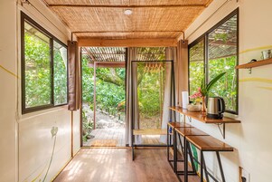 Quarto com amplas janelas, teto de madeira e uma pequena mesa com chaleira e flores.