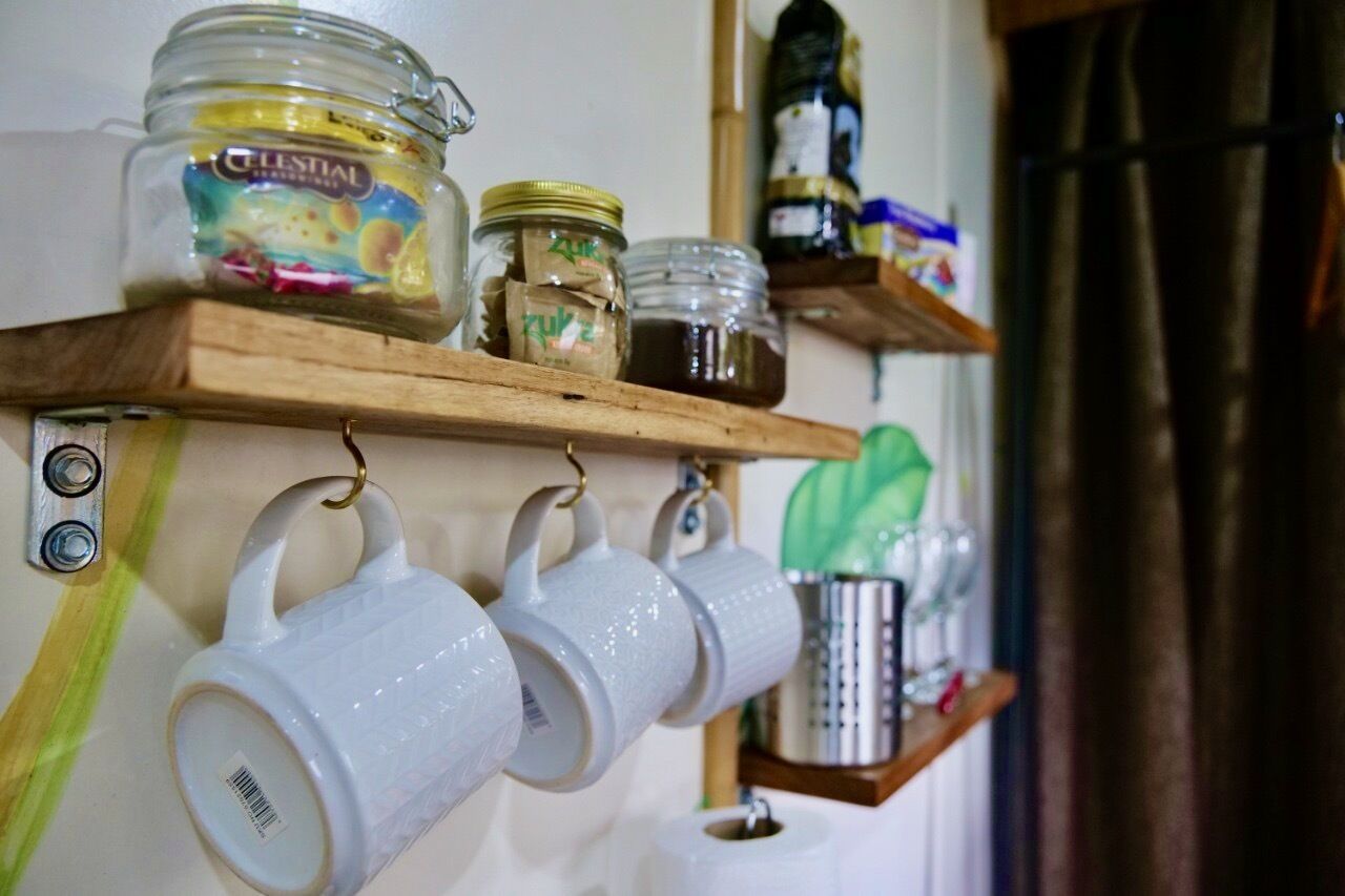A kitchen shelf with jars of condiments and hanging white mugs.