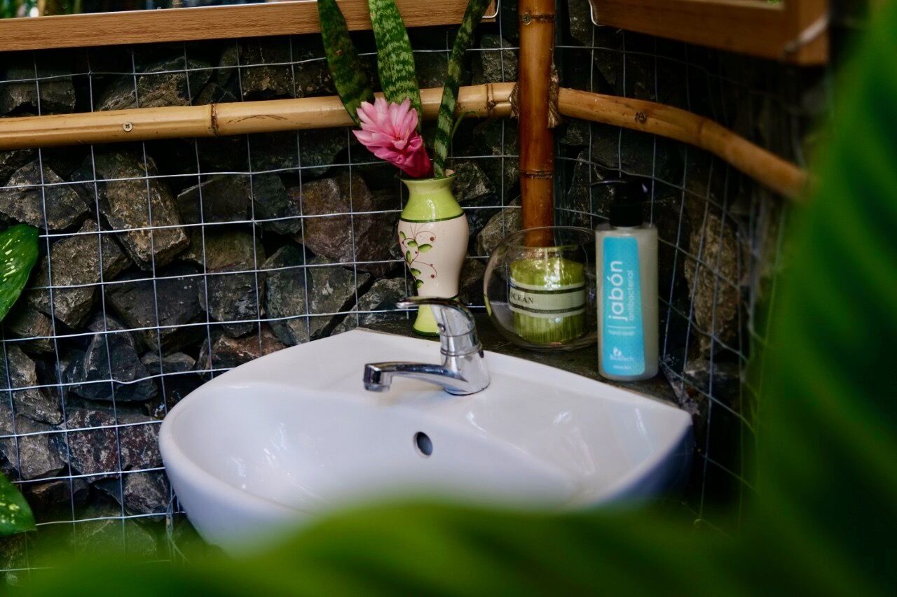 A bathroom with a white sink, a bamboo shower rod, a vase with pink flowers, and a bottle of soap.