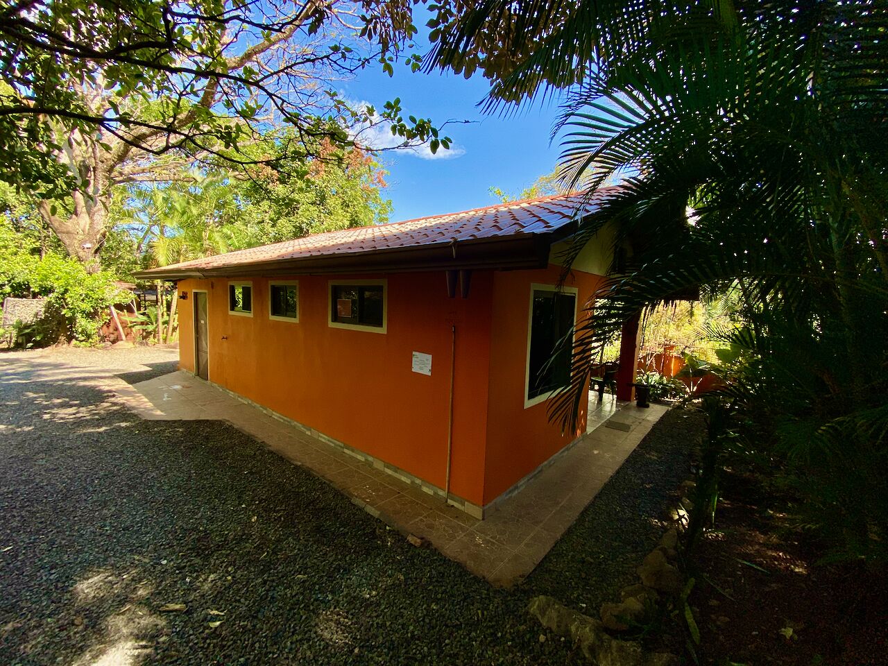Um pequeno edifício térreo com telhado vermelho e paredes bege, cercado por vegetação densa.