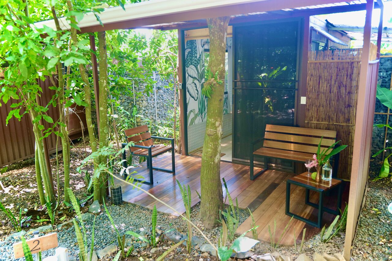 A wooden deck with a bench and a small table, surrounded by greenery and a bamboo fence.