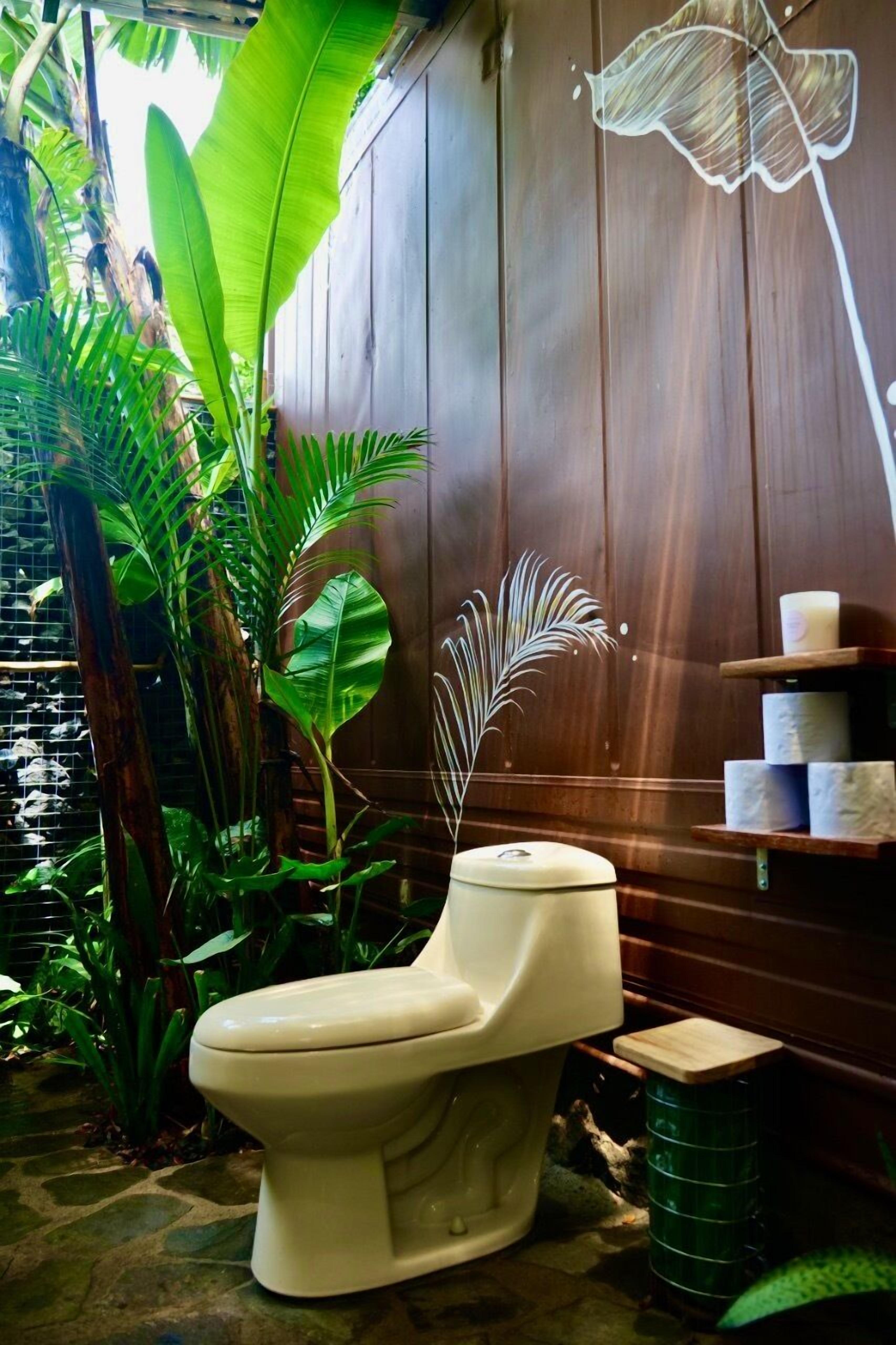 Des toilettes dans une salle de bain dont un des murs est en bois, le tout entouré de plantes tropicales.