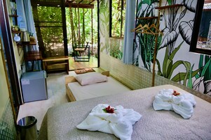 Quarto com duas camas, uma mesa pequena, uma cadeira, janela com vista para vegetação e parede com um mural de plantas.