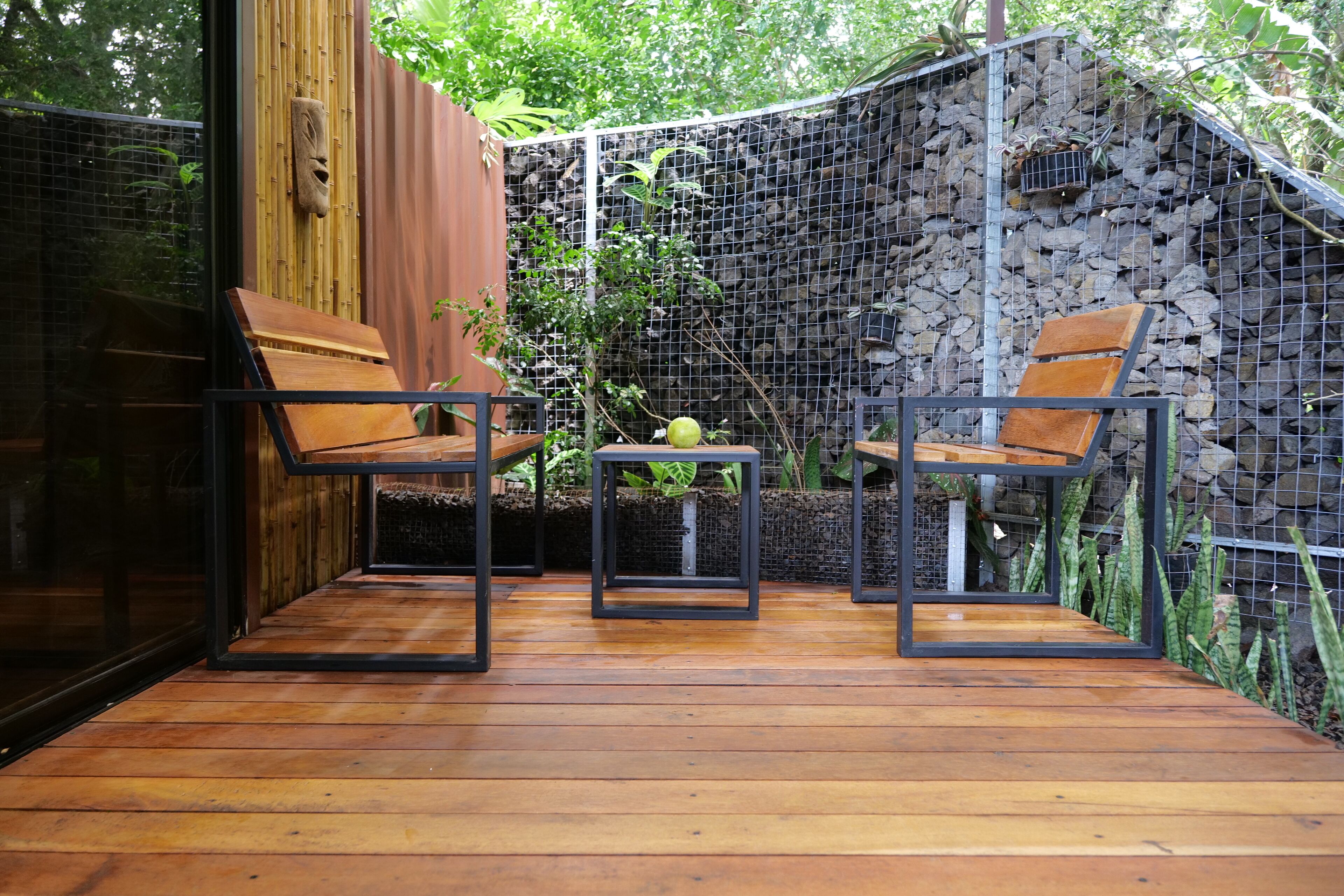 Een houten terras met twee stoelen en een kleine tafel, omgeven door een bamboe hek en een stenen muur.