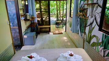 A room with a bed, a small table, and a chair. There is a window with a view of trees and a door leading outside.