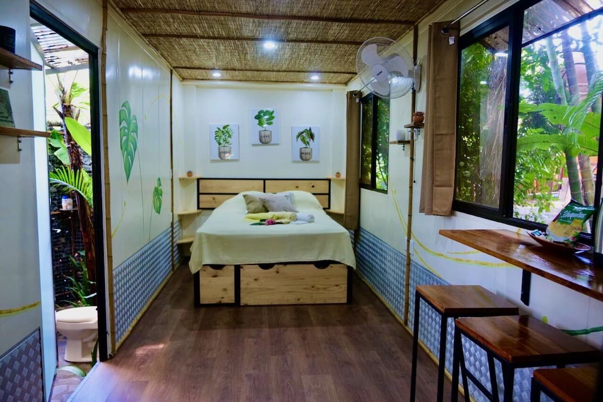 Quarto compacto com cama, ventilador, janela com vista para áreas verdes e uma pequena mesa.