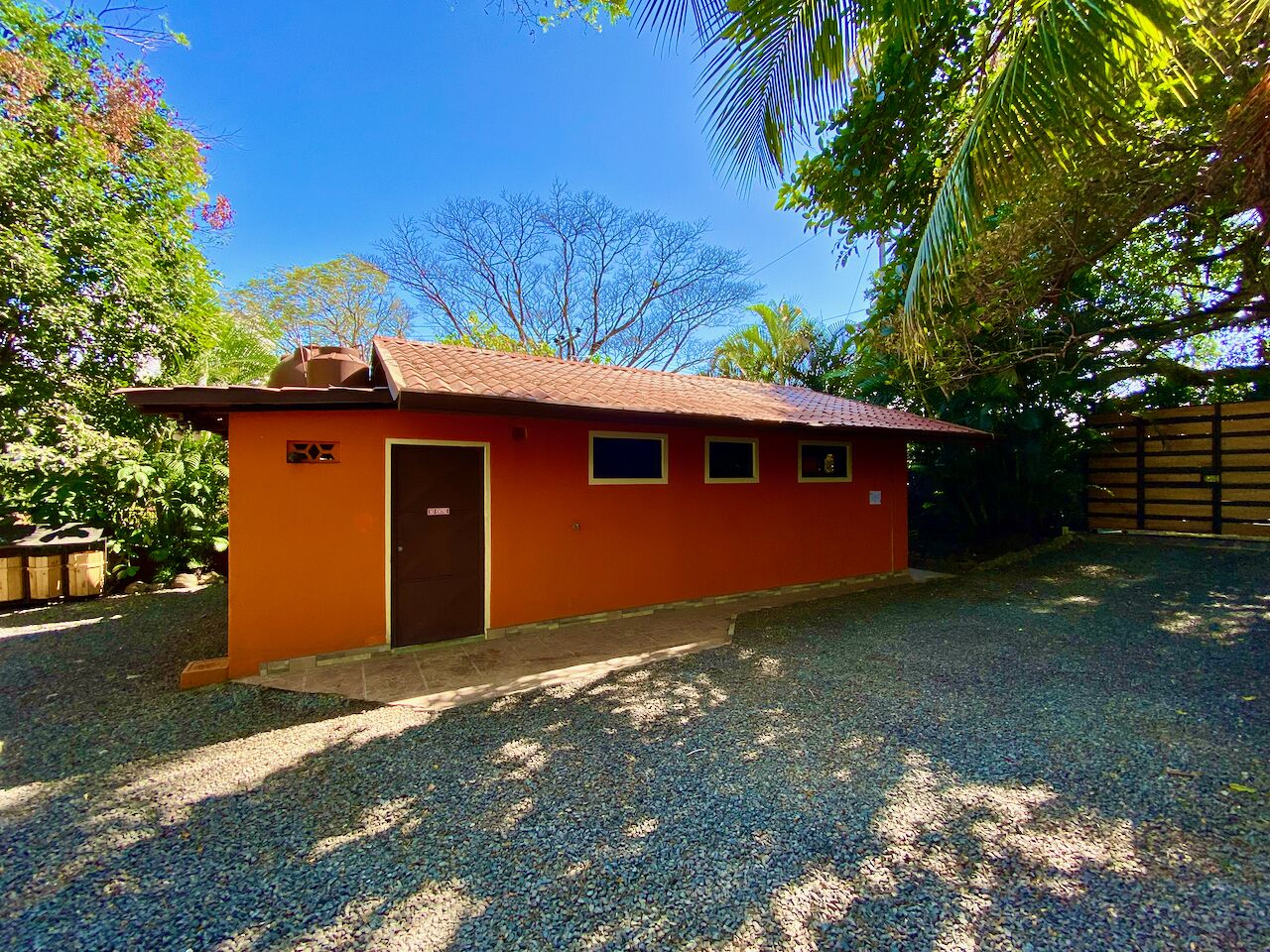 A small, single-story building with a red exterior and a brown door. It has a gravel driveway and is surrounded by trees and vegetation.