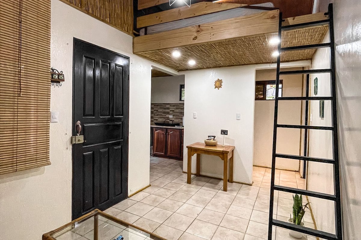 Um quarto espaçoso com uma mesa de vidro, uma porta preta, uma escada de madeira e uma pequena mesa com uma planta.