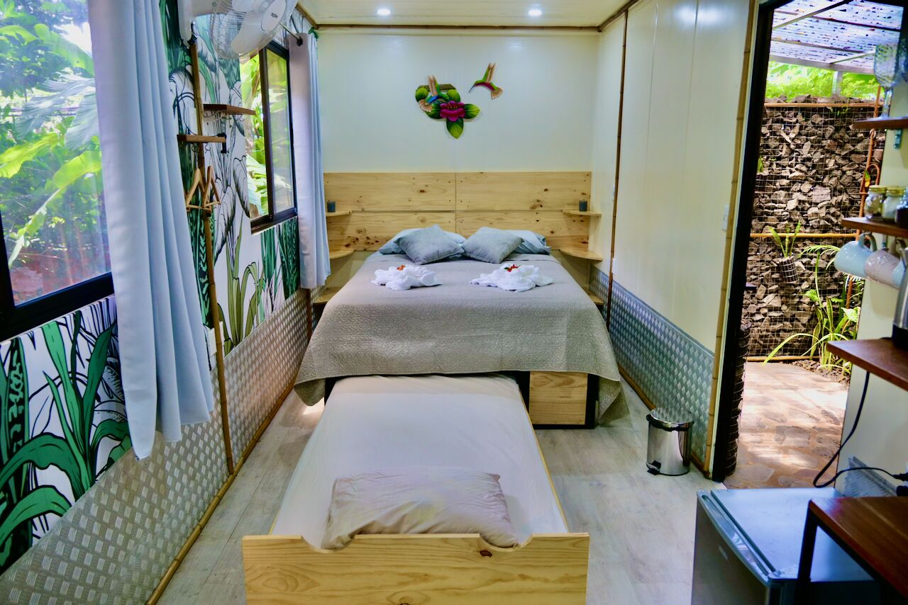 A compact bedroom with a single bed, a small bedside table, a wall-mounted fan, and a view of greenery outside.