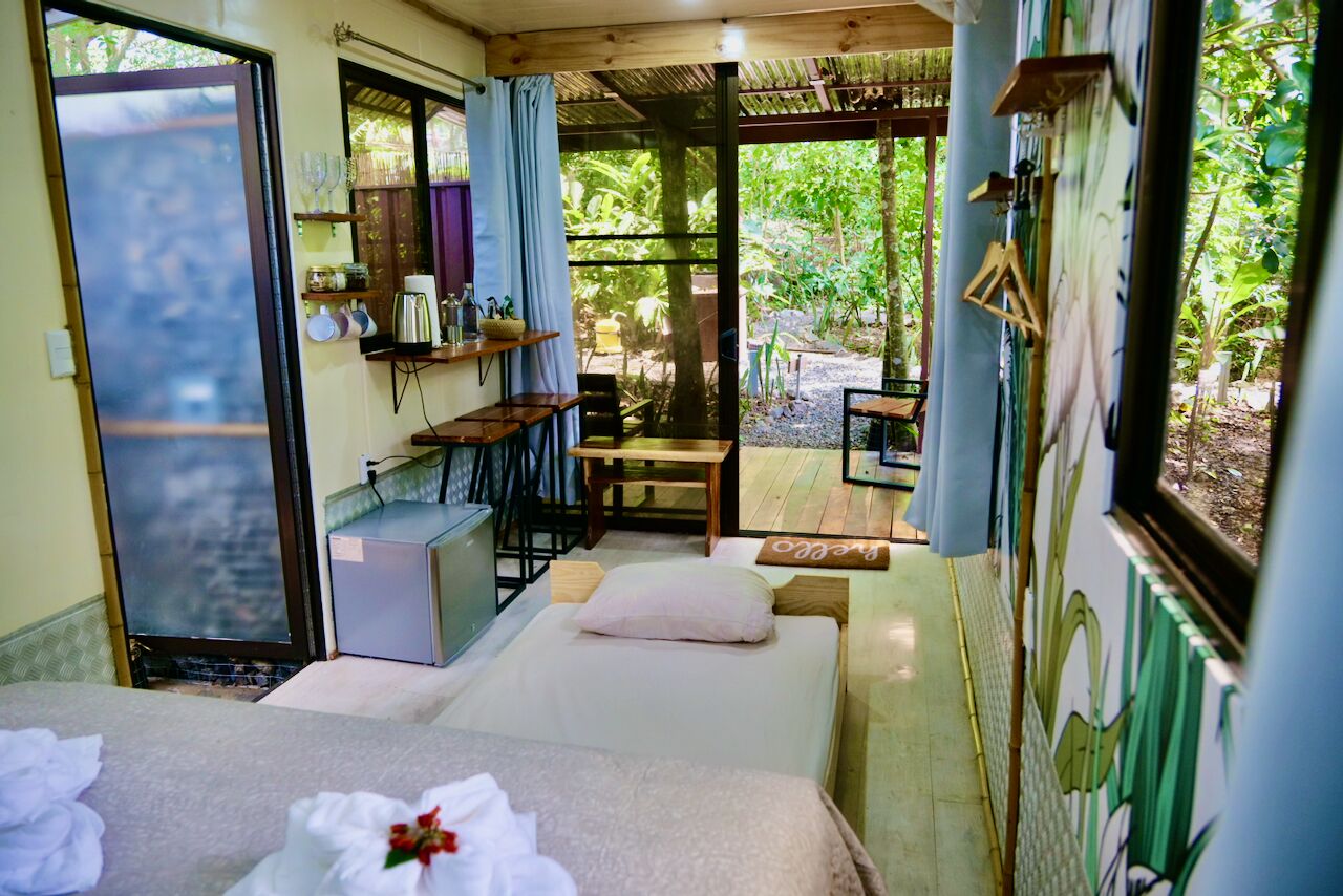 A room with a bed, a small table, and a chair, a window with a view of greenery, and a shelf with items.