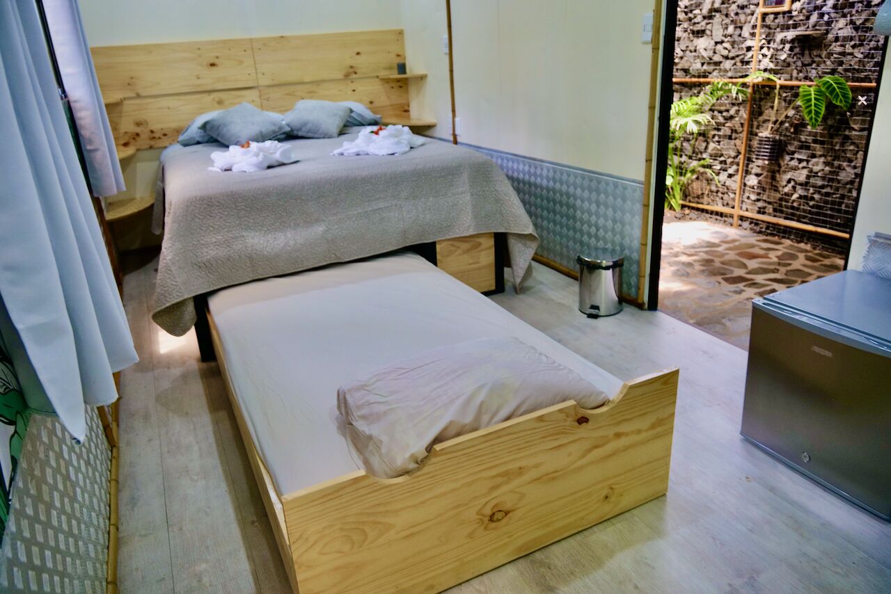 A bedroom with a wooden bed, a smaller bed, a nightstand, and a stone wall with plants.