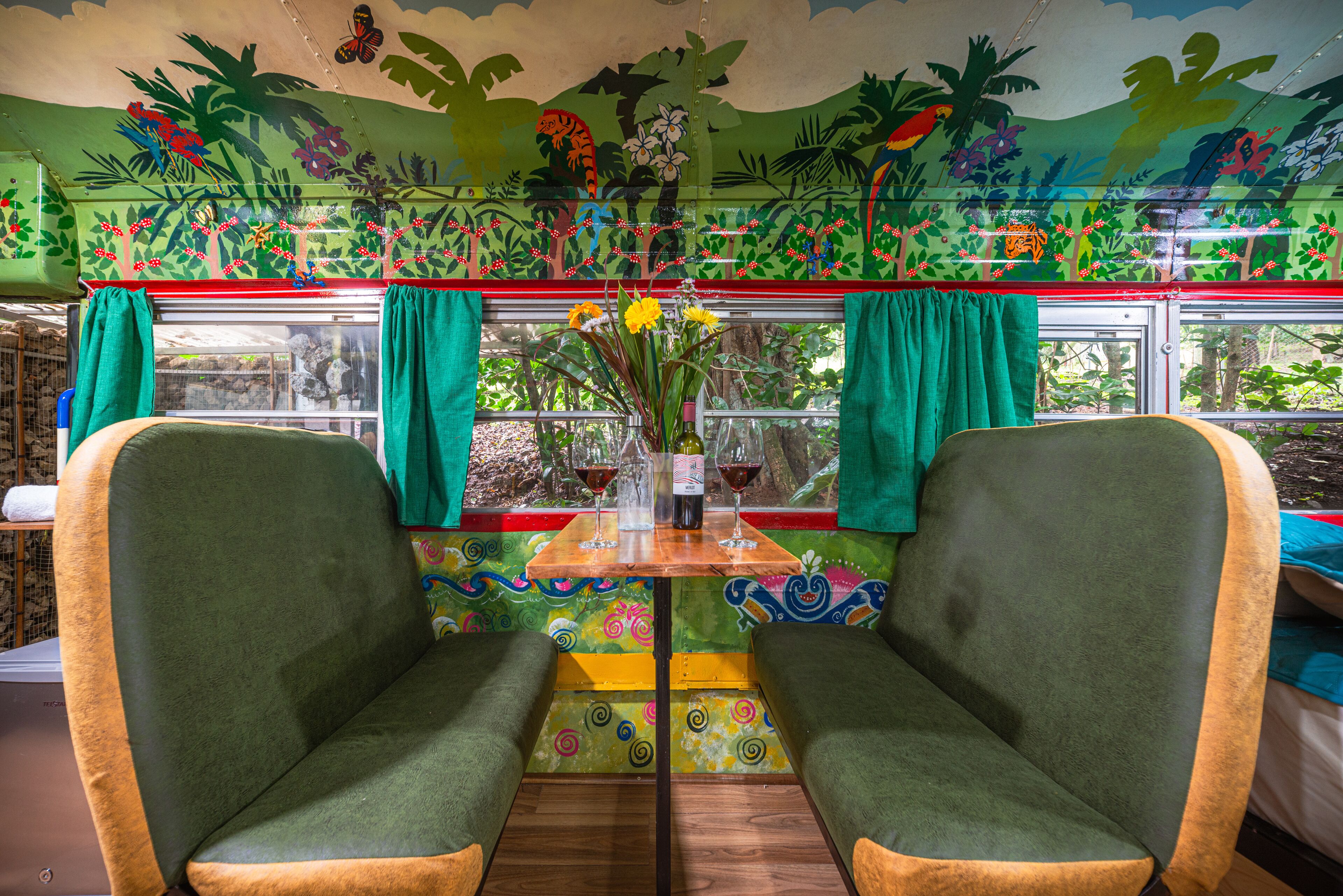 A vibrant room with a mural, a table with a vase and wine glasses, and two green upholstered chairs.