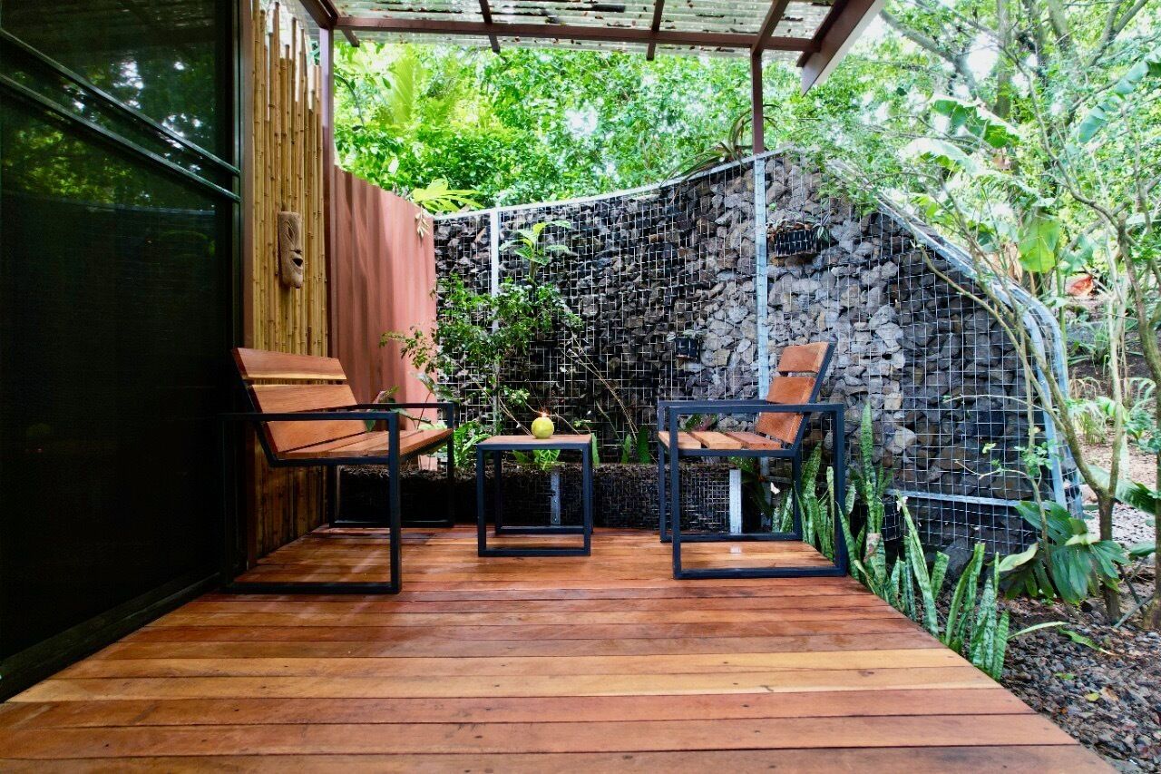 Una terraza de madera con dos sillas y una pequeña mesa, rodeada por un muro de piedra y abundante vegetación.
