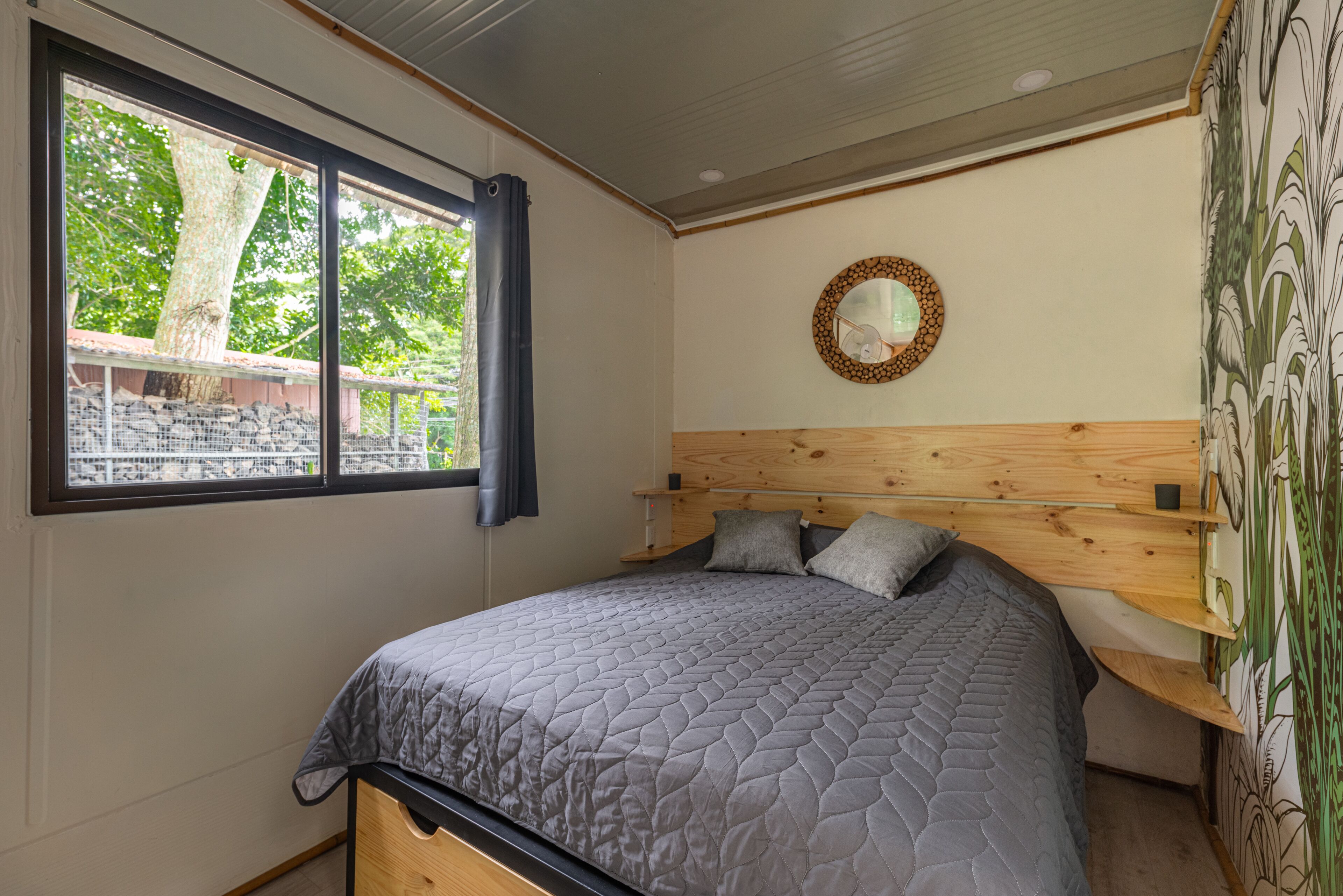 A bedroom with a large bed, a wooden headboard, a window with a view of trees, and a wall-mounted mirror.