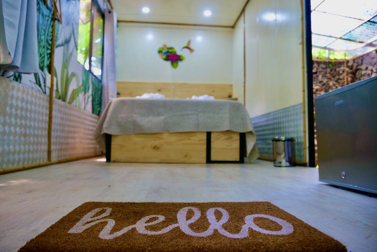 A room with a bed, a 'hello' mat, a wall with a floral decoration, and a window with curtains.