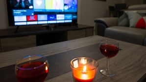 A wooden table with two glasses of red wine and a lit candle. A television is mounted on the wall in the background.