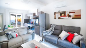 A modern living room with a grey sofa, a beige sofa, and a coffee table. There is a kitchen area with a grey refrigerator and a coffee machine. A painting is on the wall, and a window is visible.