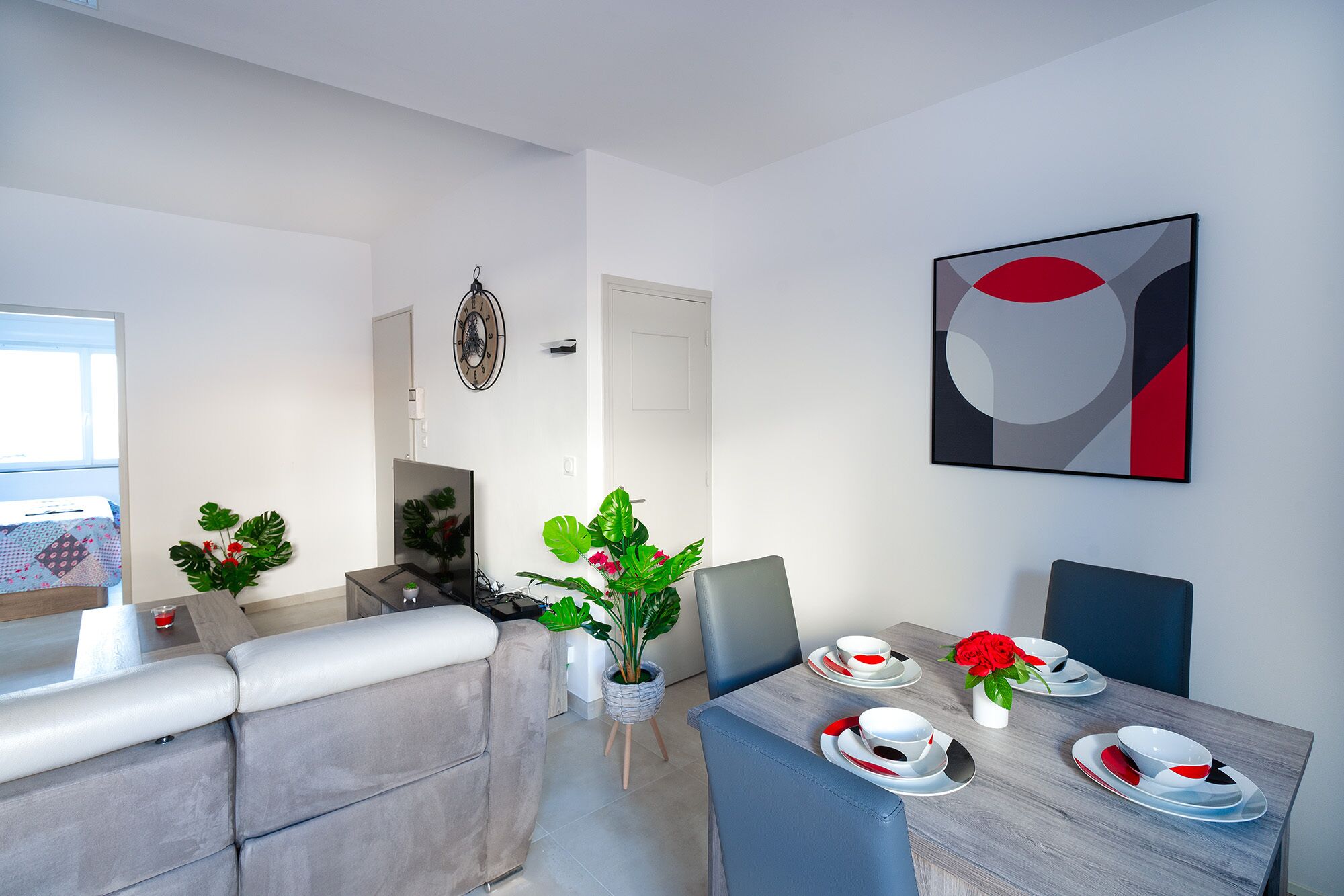 A modern dining area with a wooden table set for four, surrounded by grey upholstered chairs. A large abstract painting adorns the wall, and a potted plant adds a touch of greenery.