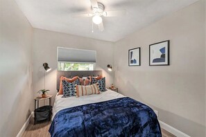 Un dormitorio con una cama, un ventilador de techo, una ventana y dos cuadros en la pared.