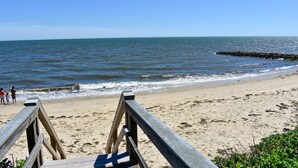 Een houten vlonder loopt naar een zandstrand, met een rotsenrijke zeebaan in de verte.