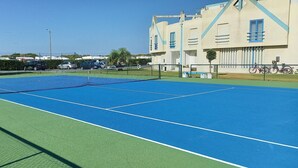 An outdoor tennis court with a net, surrounded by a fence, and adjacent to a building with blue windows and a white door.