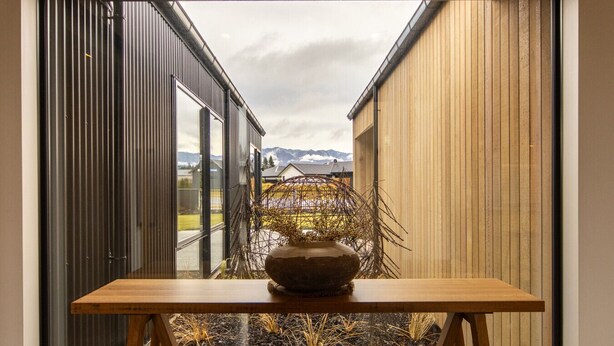 Blick durch ein Fenster. Im Vordergrund ein Tisch und eine Vase, im Hintergrund ein Holzbau mit Bergblick.