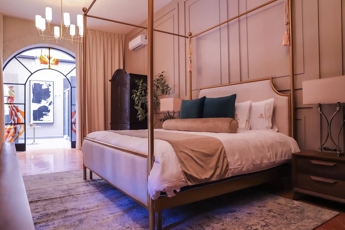 A four-poster bed with a beige bedspread and pillows, a wooden nightstand with a lamp, and a large window with curtains.