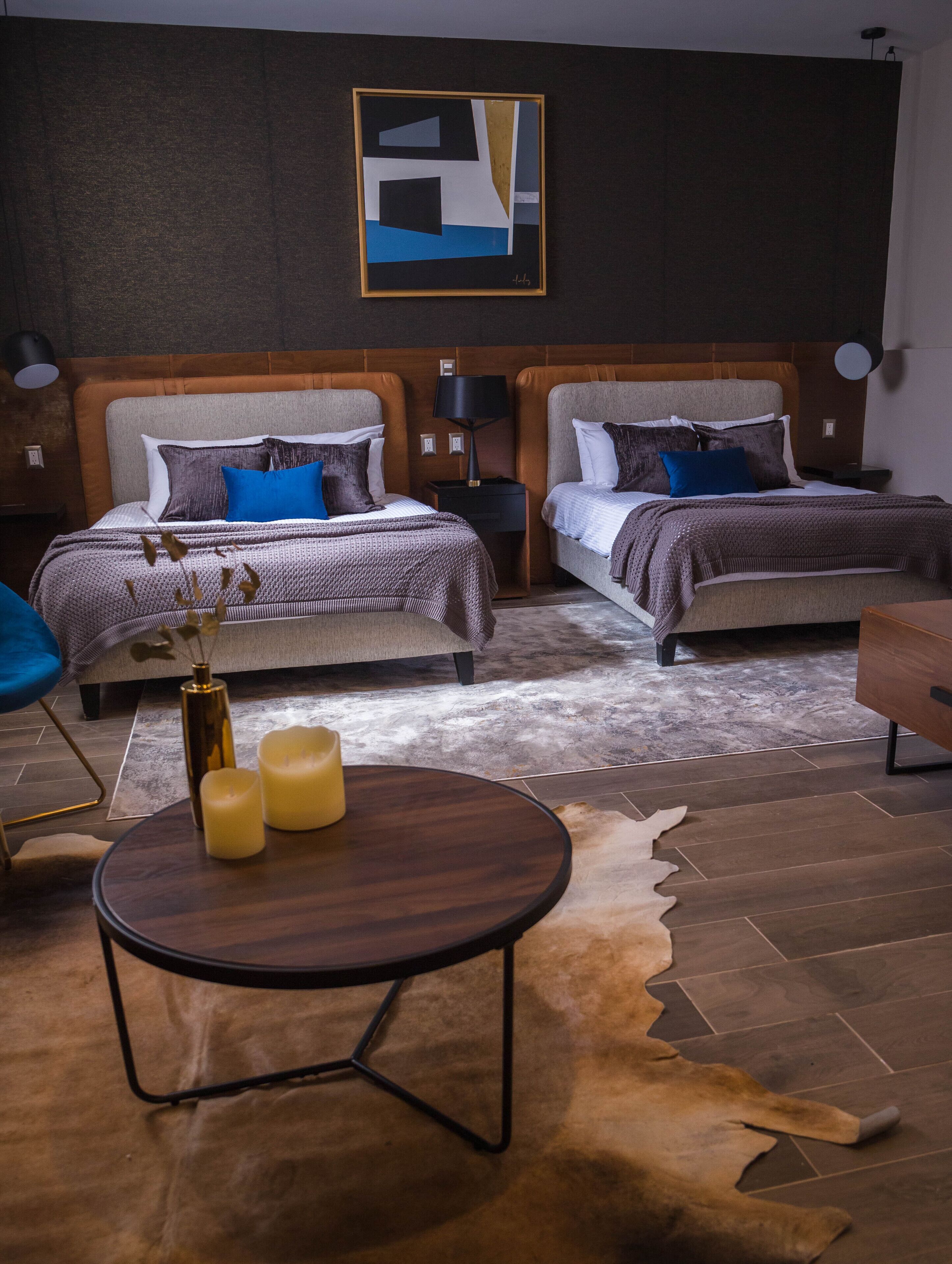 A modern hotel room with two beds, a round wooden table with candles, a cowhide rug, and a contemporary abstract painting on the wall.