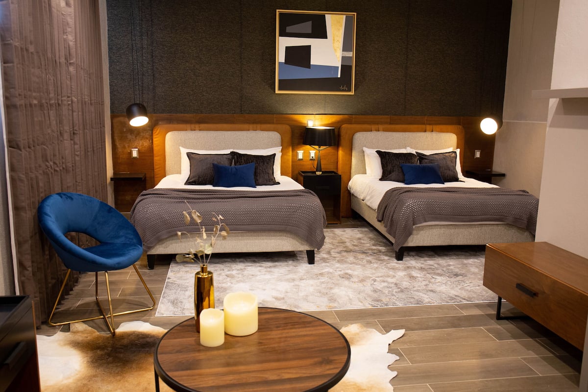 A modern hotel room with two beds, a blue chair, a wooden side table, and a rug.