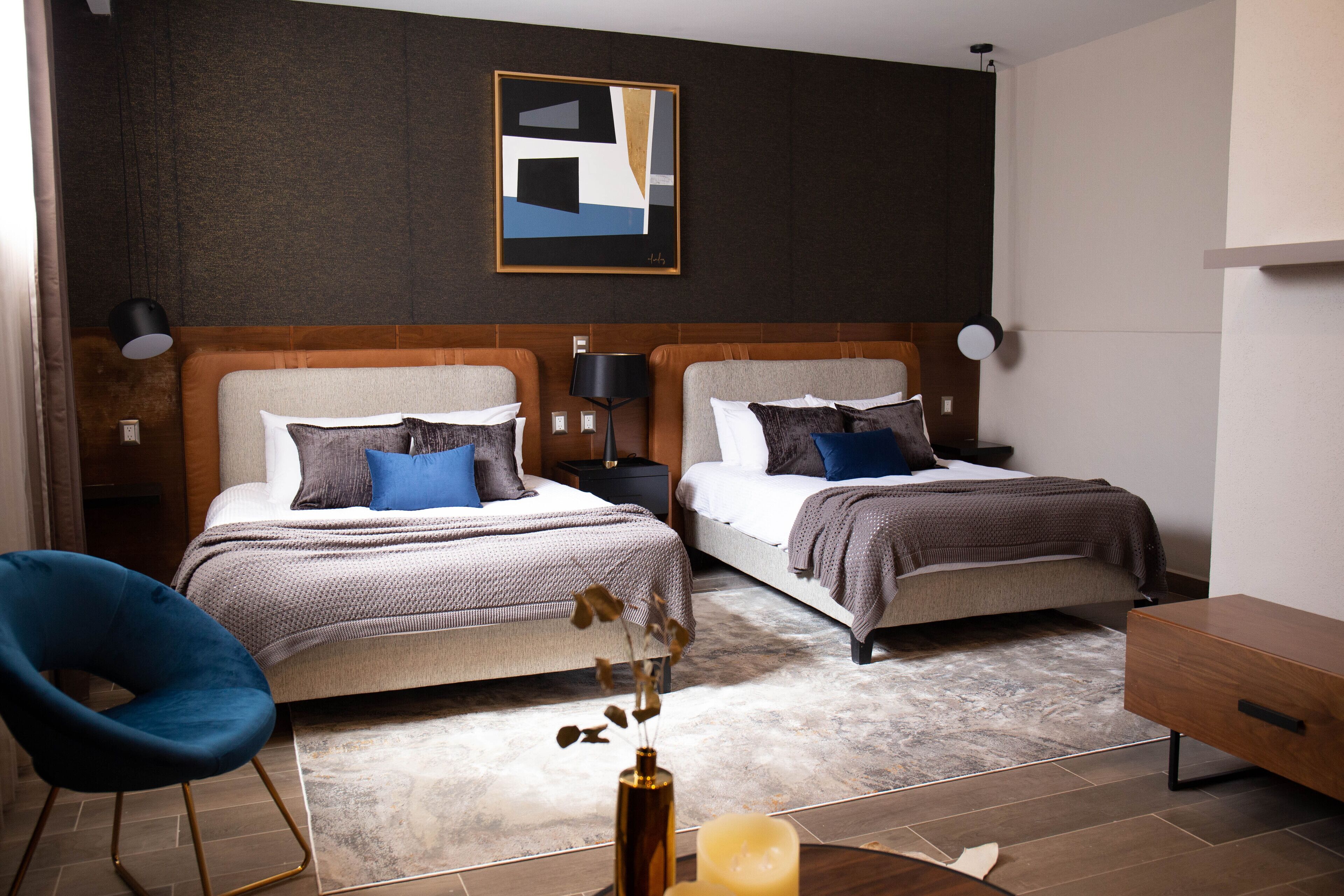 A modern hotel room with two beds, a blue chair, a wooden side table, and a framed abstract artwork on the wall.