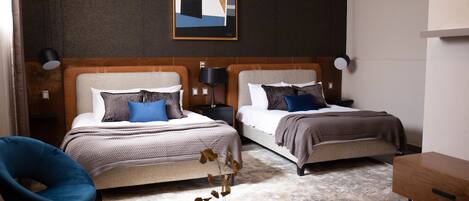 A modern hotel room with two beds, a blue chair, a wooden side table, and a framed abstract artwork on the wall.