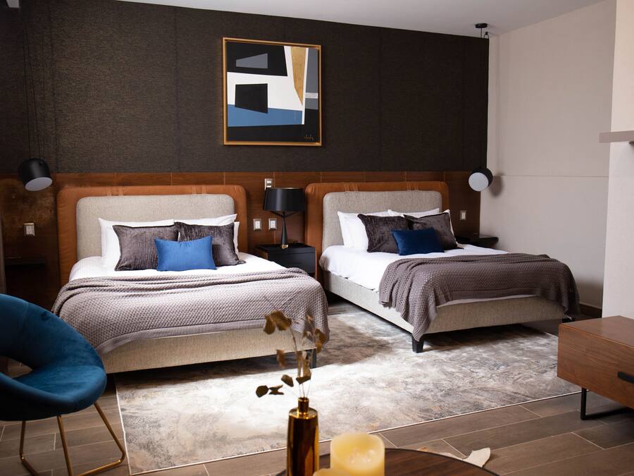 A modern hotel room with two beds, a blue chair, a wooden side table, and a framed abstract artwork on the wall.