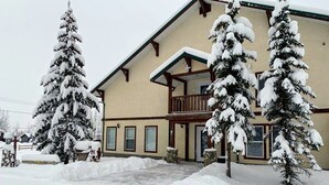 A two-story building with a balcony, snow-covered trees, and a snow-covered driveway.