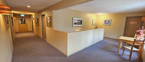 A hotel reception area with a reception desk, framed pictures on the wall, a wooden chair, and a small table with a vase of flowers.